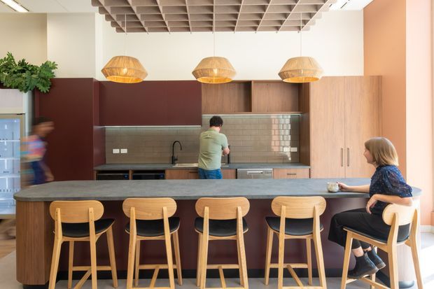 A contemporary commercial-style kitchen and communal dining area with a long dark stone island, five timber bar stools, timber cabinetry, dark burgundy panels, grey-green tiled splashback, and three woven pendant lights. The space includes a visible sink and appliances, with a distinctive exposed grid ceiling and people using the kitchen and island.