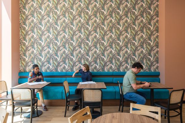 A bright contemporary café or casual dining space with a long turquoise upholstered banquette set against a bold botanical-patterned wallpaper feature wall. Small wood tables, mixed cane and timber chairs, and several people using the seating create an inviting, colourful hospitality interior. Warm terracotta-toned side walls and light timber flooring complement the vivid blue-green upholstery.