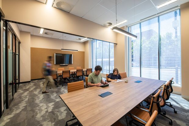 Bright contemporary commercial meeting and collaborative workspace with a large timber conference table, surrounding office chairs, a smaller meeting area with wall-mounted display, glass-partitioned rooms, floor-to-ceiling windows, suspended linear lighting, and patterned carpet flooring.