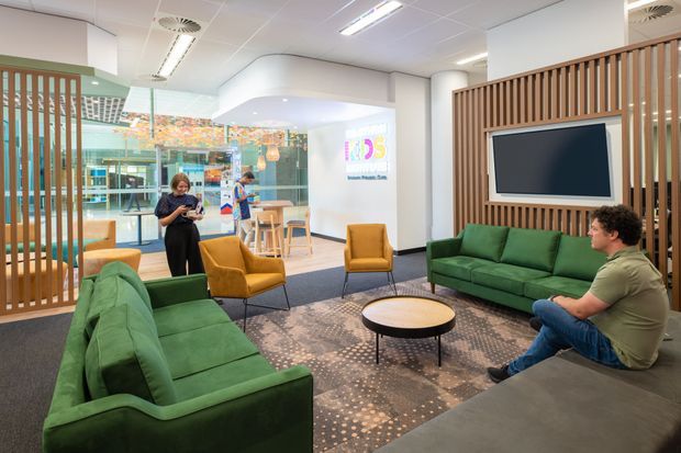 Bright contemporary commercial lounge and reception area with green sofas, mustard armchairs, a round coffee table, patterned carpet, timber slatted room dividers, and a wall-mounted screen. Glazed meeting or breakout spaces and a café-style counter area are visible in the background.
