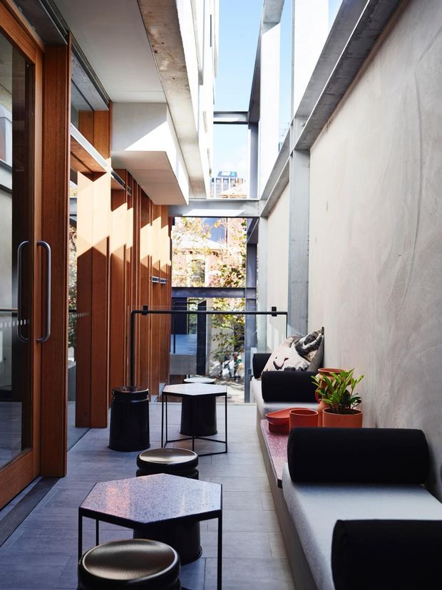 A narrow, elongated contemporary interior lounge or passage with full-height glazing and timber doors on the left, a smooth concrete wall on the right, and a series of black metal-framed coffee tables and stools. Built-in or linear seating runs along the concrete wall, accompanied by potted plants and strong natural daylight. The space opens toward a glazed courtyard or urban outlook at the far end.