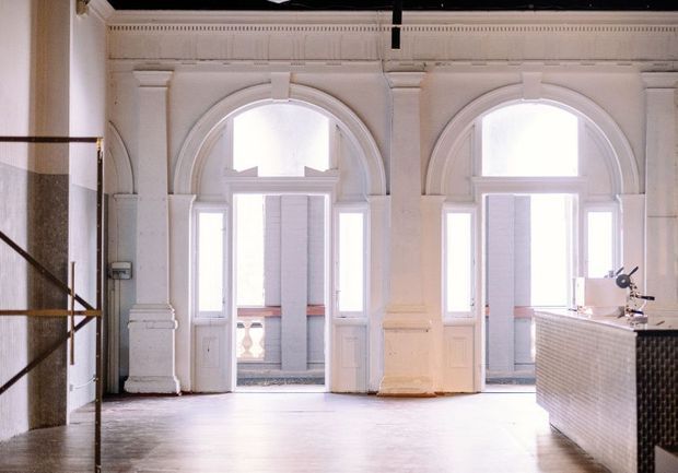 Bright, largely empty interior with two tall arched French doors and windows, ornate white classical wall mouldings and pilasters, and a dark tiled reception or counter structure at right. The space has a polished, gallery-like character with exposed architectural detailing and abundant natural light.