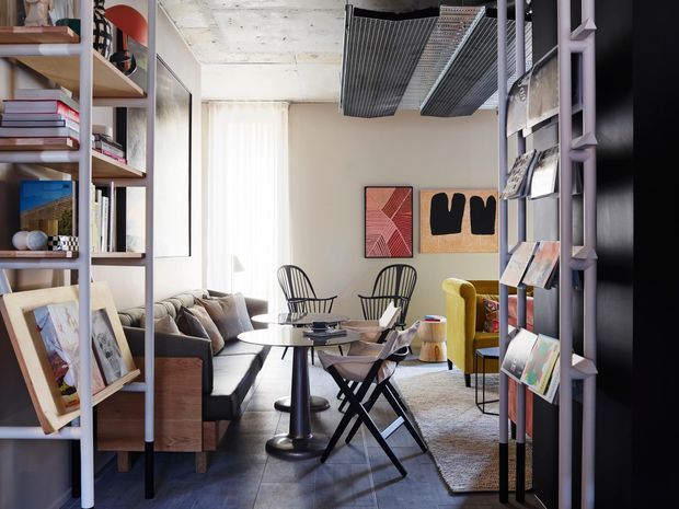 A stylish compact living room or lounge with an eclectic, art-led interior. Open shelving and magazine display racks frame the view, while a sofa, round dining or occasional table, chairs, a yellow armchair, framed artwork, and a textured rug create a layered, inviting space. The room features an exposed concrete ceiling, dark tiled flooring, pale walls, and prominent black and lavender-toned metal shelving.