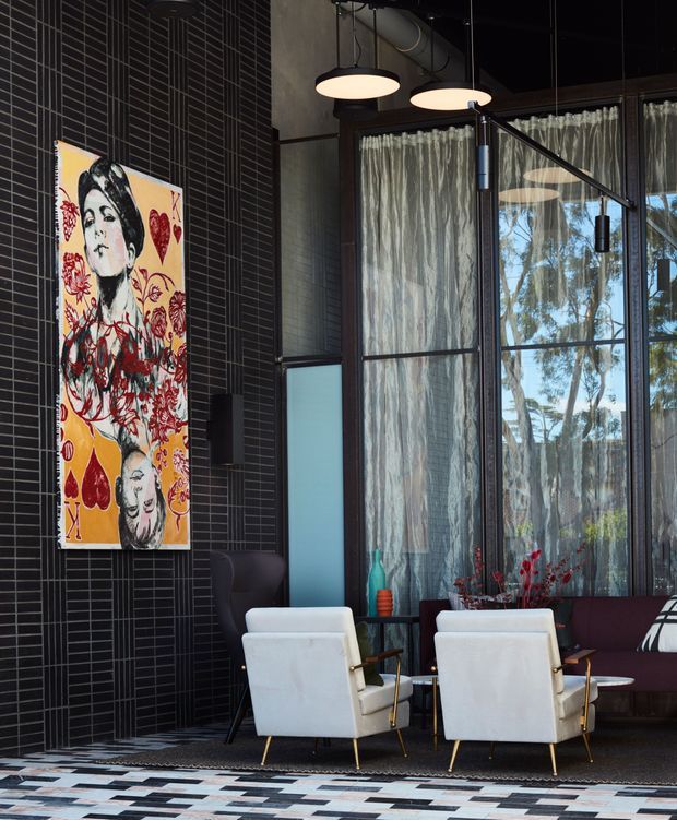 A sophisticated lounge or lobby interior with a dramatic black linear tiled feature wall, full-height industrial-style glazed partitions with sheer curtains, exposed services and pendant lighting. Two white mid-century-style armchairs face a dark upholstered sofa, with a colourful figurative artwork and patterned geometric floor adding bold accents.