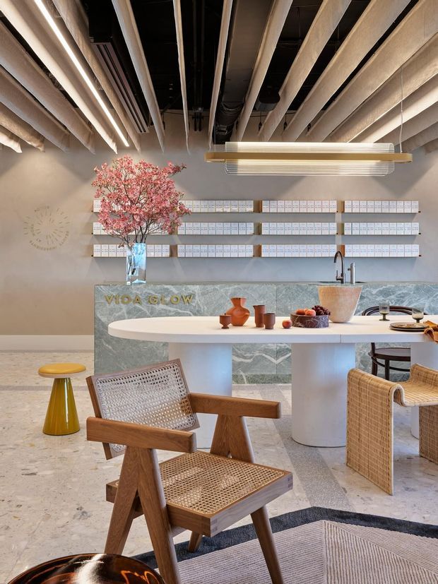 A warmly lit contemporary commercial interior featuring a reception or hospitality counter with a pale green stone front, white rounded island-style table, woven cane and timber seating, and a decorative pink flowering tree. The exposed ceiling reveals ducts, services, and linear lighting, while wall-mounted shelving or display panels sit behind the counter. The space has a refined, organic, softly colourful aesthetic.