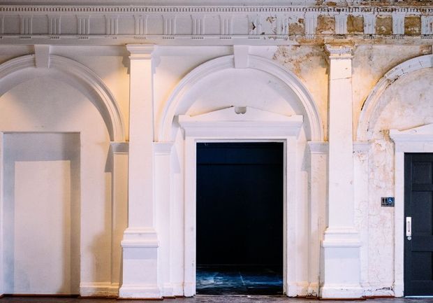 A close-up architectural interior view of an ornate, weathered white plaster facade with repeated arched openings, pilasters, classical mouldings, and decorative cornices. The central opening leads into a dark room, while a black door is visible at right. Peeling, stained plaster contrasts with carefully detailed classical ornamentation.