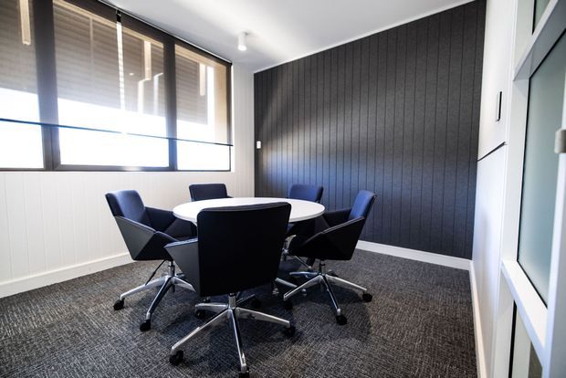 A compact contemporary meeting room with a round white table surrounded by black upholstered swivel office chairs. The room has dark vertical wall panelling, white lower wall panelling, large black-framed windows with blinds, carpet flooring, and bright recessed or track-style ceiling lighting.