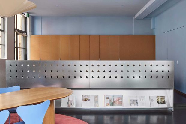 A contemporary interior reception or communal workspace with a long built-in timber storage wall, perforated metal screen, integrated display shelving, and a curved timber table with blue chairs. Tall industrial-style windows line the left side, while pale blue walls, dark polished flooring, and a red area rug create a refined, slightly playful atmosphere.