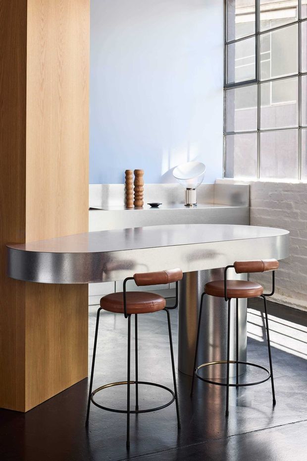 A bright, compact contemporary interior featuring a polished stainless-steel island or bar counter with cylindrical supports and two leather-topped bar stools. A warm light-oak vertical cabinet or wall panel frames the left side, while a large black-framed industrial-style window and exposed white-painted brick wall occupy the right. The space has a dark polished floor, pale blue-white walls, and minimal decorative objects, creating a refined modern loft-like atmosphere.