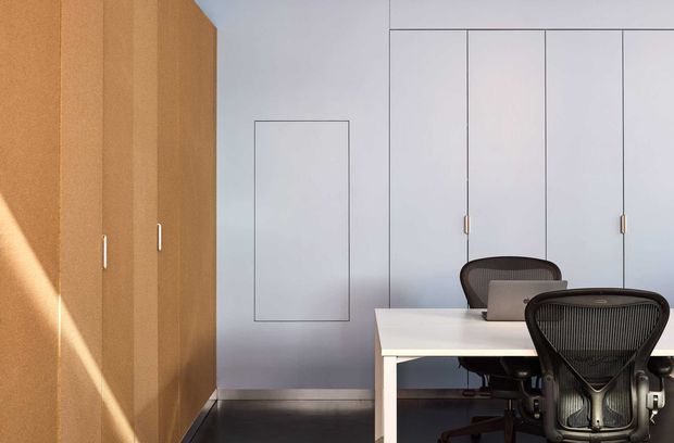 Minimalist contemporary home office with a white desk and ergonomic mesh office chairs, set against pale blue-grey built-in cabinetry or wall panelling. A warm timber built-in storage wall occupies the left side, with slim vertical handles and strong sunlight creating a diagonal highlight. The space has clean lines, restrained detailing, and a dark floor.