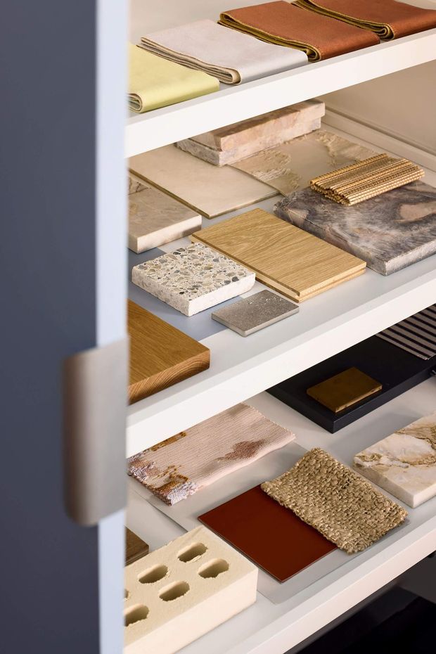 Close-up of an open sample storage cabinet displaying an organised collection of interior and architectural material samples, including folded textiles, stone, terrazzo, timber, tile, metal, leather-like sheets and woven surfaces. The shelves and cabinet are white, with a dark grey-blue side panel and handle visible in the foreground. The image is a materials library or design showroom display rather than a finished room.