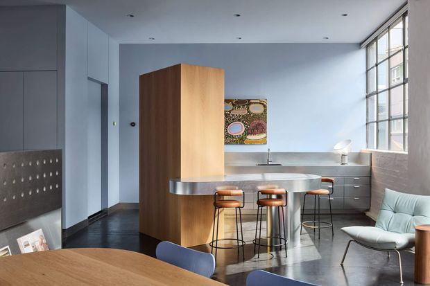 A compact, open-plan contemporary kitchen and dining area with a curved stainless-steel island and raised breakfast counter, three bar stools, dark fixed cabinetry, and a tall timber-clad storage volume. The room has pale blue-grey walls, polished dark flooring, exposed brickwork along the right wall, a large industrial-style steel-framed window, recessed ceiling lights, and a light lounge chair. A timber dining table occupies the foreground.