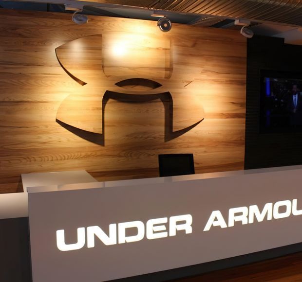 A dramatic reception or showroom counter with illuminated branding, set against a warm timber feature wall displaying a large sculptural logo. Track lighting highlights the wood wall, while a dark media screen and dark ceiling elements add contrast.