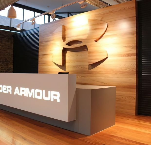 A modern commercial reception area featuring an illuminated Under Armour logo mounted on a large warm timber feature wall. A white angular reception desk and adjacent pedestal sit on a timber floor, with black metal screening, exposed masonry, expansive glazing, and contemporary pendant lighting contributing to an industrial-modern workplace aesthetic.