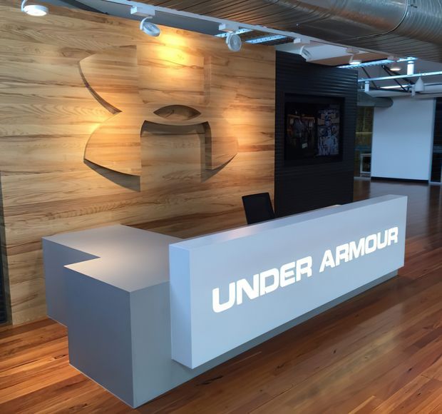 A branded commercial reception area featuring a large illuminated Under Armour reception desk, warm horizontal timber wall panelling with an inset sculptural logo, timber flooring, exposed ductwork, and track lighting. A dark feature wall and office-style glazing are visible in the background.