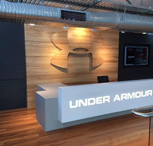 Interior view of an Under Armour reception area in a commercial workplace or showroom. The space features a prominent illuminated white reception desk, a large carved timber feature wall with the brand logo, dark ribbed wall panels, exposed silver HVAC ductwork, and timber flooring. The lighting is warm and focused, creating a polished branded entry environment.