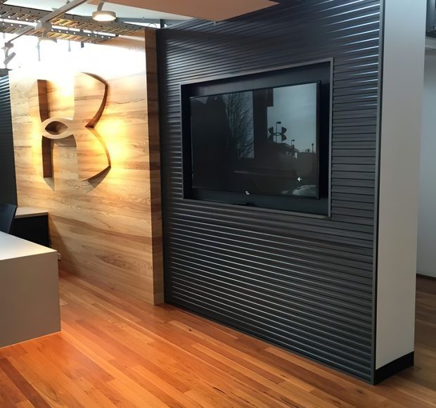 A contemporary interior feature wall with a built-in television set into dark horizontal ribbed panelling. Adjacent timber wall panelling features a large sculptural cut-out logo or artwork, while warm timber flooring, a white counter or cabinet, exposed ceiling elements, and pendant lighting complete the space.