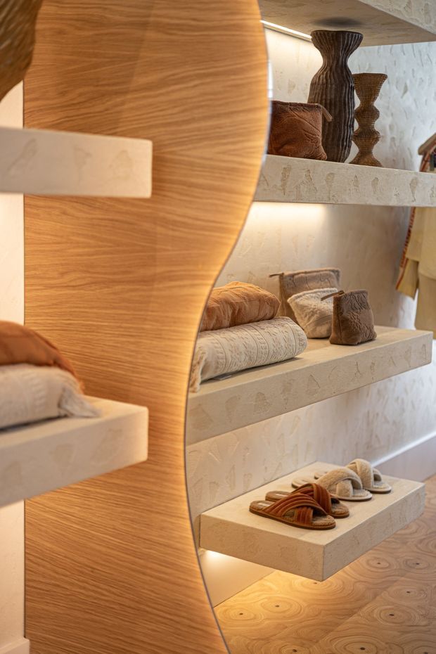 Close-up interior view of a warm, sculptural retail display or dressing area. Curved light timber joinery forms a prominent vertical divider, while illuminated floating shelves display folded textiles, cushions, decorative vessels, and sandals. The shelves and adjacent wall feature a textured pale stone or plaster finish, complemented by warm integrated lighting and a timber floor.