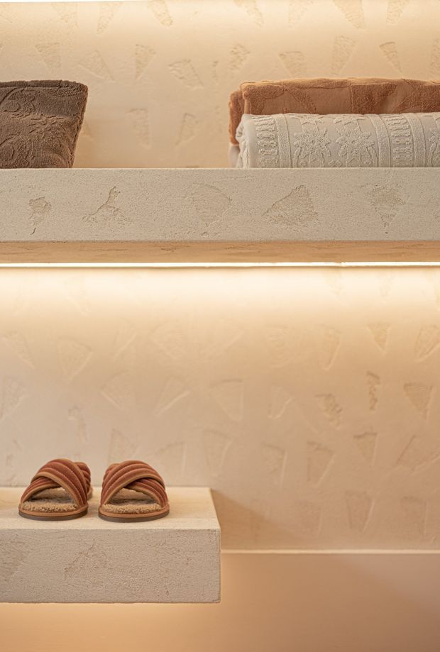 Warmly lit built-in bathroom shelving or niche with textured plaster finishes, rolled towels stored on the upper shelf, and sandals displayed on a lower floating ledge. The monochromatic beige and terracotta palette creates a calm, tactile spa-like atmosphere.