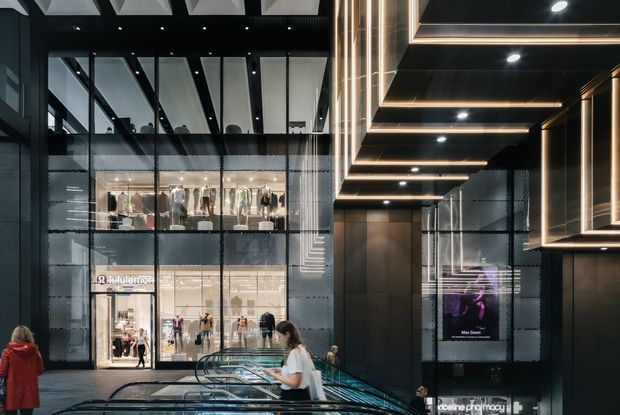 A dramatic contemporary commercial retail interior with a dark, highly detailed architectural fit-out. The image looks across a multi-level shopping centre or fashion retail space, featuring extensive floor-to-ceiling glazing, visible shopfronts, escalators, black ribbed and patterned wall surfaces, dark timber-toned panels, and layered recessed ceiling lighting with warm linear illumination. Bright retail displays and signage are visible behind the glazing, while shoppers and escalators provide scale.