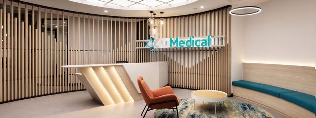 A contemporary medical reception and waiting area featuring a curved reception desk, vertical timber slat screening, illuminated desk panels, integrated bench seating, a round coffee table, and a prominent AD Medical sign. The space has a warm neutral palette with teal and orange accents and a patterned rug.
