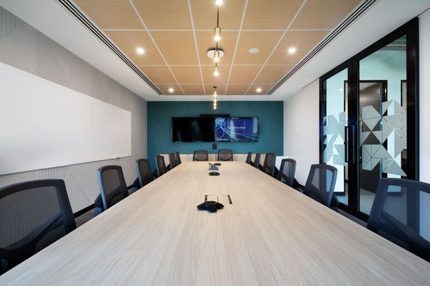 A bright contemporary commercial meeting room with a long light-wood conference table surrounded by black mesh office chairs. The room has a teal feature wall with a wall-mounted display, white acoustic wall panels, a warm wood acoustic ceiling grid with recessed lighting, and black-framed glazed doors featuring geometric privacy decals.