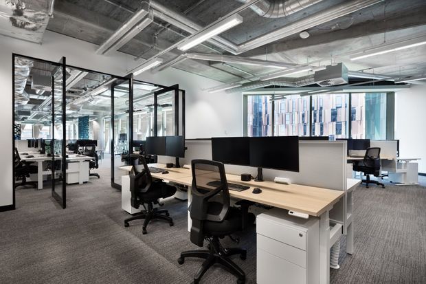 Contemporary open-plan commercial office interior with rows of light-wood workstations, black ergonomic office chairs, computer monitors, white drawer units, and extensive glass-partitioned meeting or work areas. The exposed concrete ceiling and visible mechanical ductwork create an industrial character, while large windows provide abundant daylight.