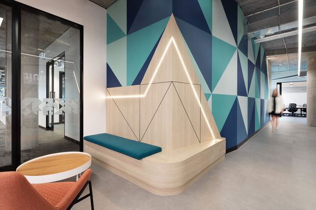 A contemporary commercial interior lobby or reception area with a sculptural built-in timber seating nook. The feature joinery has pale wood panelling, teal upholstered bench cushions, integrated linear LED lighting, and angular geometric detailing. A bold geometric mural in navy, teal, white, and pale blue extends along the corridor wall. Black-framed glazed meeting-room doors, exposed concrete and services, and modern pendant lighting contribute to an industrial-contemporary workplace aesthetic.