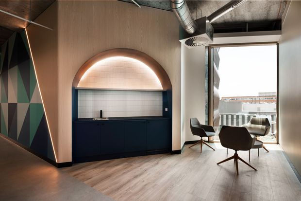 A sophisticated office lounge or meeting area with a dark navy built-in kitchenette or refreshment bar beneath a rounded, illuminated arch. The space features warm timber wall panelling and wood-look flooring, exposed industrial ductwork, geometric dark-and-green wall artwork, and mid-century-inspired lounge chairs arranged beside floor-to-ceiling glazing. The palette is moody and refined with a strong blend of contemporary, industrial, and mid-century elements.