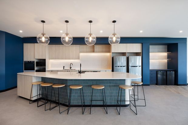 A spacious contemporary kitchen with a large curved island and integrated seating for multiple people. The island features a pale stone benchtop and vertically patterned blue-green tile cladding, while the rear cabinetry combines light timber, dark blue cabinetry, open shelving, and illuminated textured tile splashbacks. Four spherical pendant lights hang above the island, with concealed appliances and multiple refrigerators along the back wall.