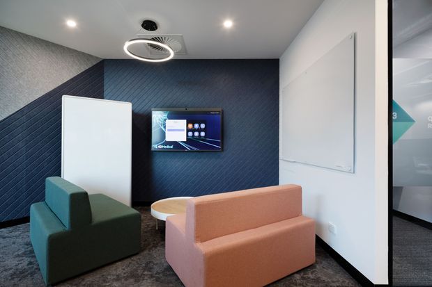 A compact contemporary waiting or breakout lounge with two upholstered sofas, a wall-mounted digital display, acoustic-style dark blue wall panels, a white presentation board, recessed ceiling lights, and a circular pendant fixture. The space appears to be a modern commercial interior, likely an office reception or collaboration area.