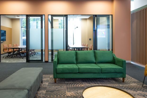A contemporary commercial office lounge and meeting area with a deep green sofa in the foreground, a low round timber coffee table, patterned rug, and a grey upholstered ottoman. Behind the lounge are black-framed glazed meeting rooms with frosted privacy panels, office tables and chairs, and a warm peach-toned wall. A vertical timber slat feature wall appears at right.