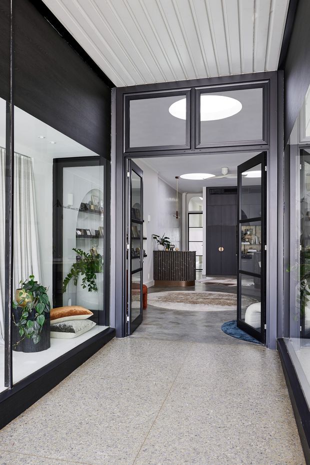 A contemporary entryway viewed from a glazed front porch or vestibule, with dark-framed glass doors opening into a bright interior. The space features polished grey terrazzo flooring, dark timber or painted wall panels and joinery, a white-lined ceiling, indoor plants, display shelving, and a curved reception-style cabinet visible beyond the entrance.