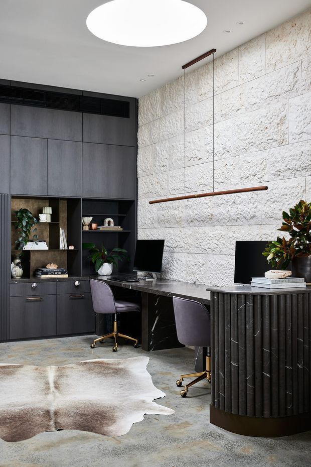 A sophisticated contemporary home office with two workstations, dark built-in cabinetry, black marble-effect desks, purple upholstered swivel chairs, and decorative shelving. The room features a dramatic rough white stone block feature wall, polished concrete-style flooring, a sculptural round ceiling light, linear pendant lighting, and a dark fluted reception or storage console. Neutral tones are accented by greenery, books, and warm timber and brass details.