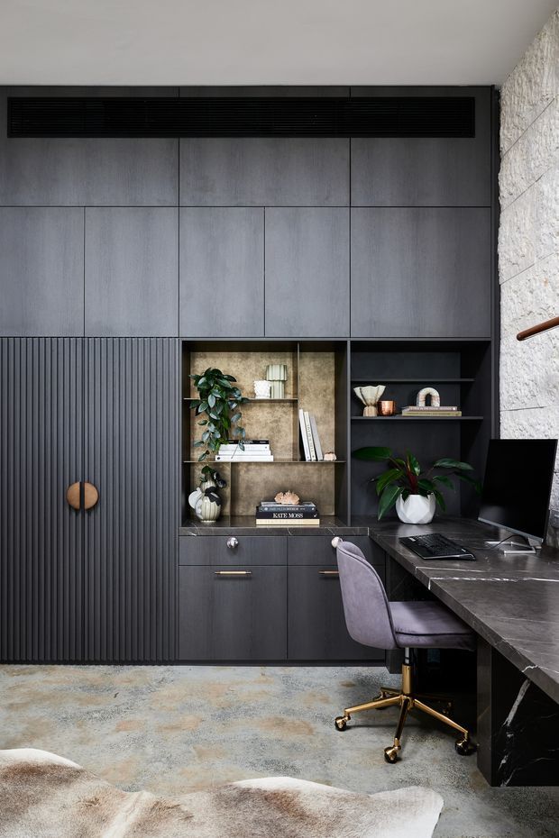 A sophisticated home office with dark built-in cabinetry, a long stone-look desk, integrated shelving, ribbed cupboard doors, and a black ventilation grille. The room features a textured pale stone wall, concrete-toned carpet, a modern upholstered office chair with brass-coloured base, indoor plants, and carefully styled shelving.