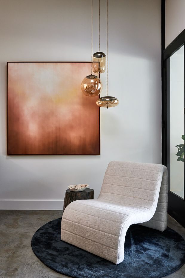 A refined interior vignette featuring a sculptural cream upholstered lounge chair on a dark round rug, set against a pale wall with a large copper-toned abstract artwork. Three suspended amber glass pendant lights hang beside the artwork, while a dark-framed glazed opening and a small organic side table complete the composition.