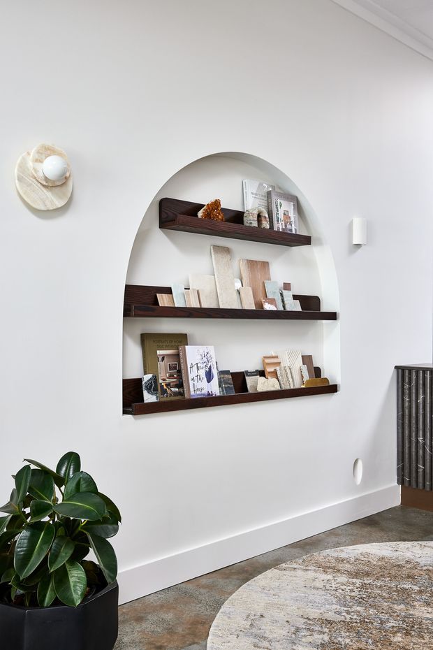 A bright, refined interior wall featuring a recessed arched alcove with three dark timber display shelves holding books, ceramics, and small decorative objects. White plaster walls, minimalist wall lighting, a large leafy plant in a black planter, a textured neutral rug, and a dark vertical cabinet or radiator cover complete the scene.