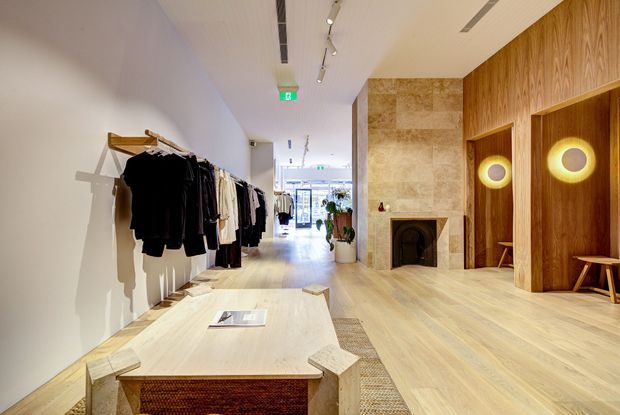 A refined retail clothing boutique with a long, bright circulation space. Garments hang along the left wall, while the right side features warm timber wall panelling, recessed fitting-room or display niches with circular illuminated mirrors, a stone-clad fireplace, and light timber flooring. A central low timber display table and track lighting contribute to the contemporary, welcoming interior.