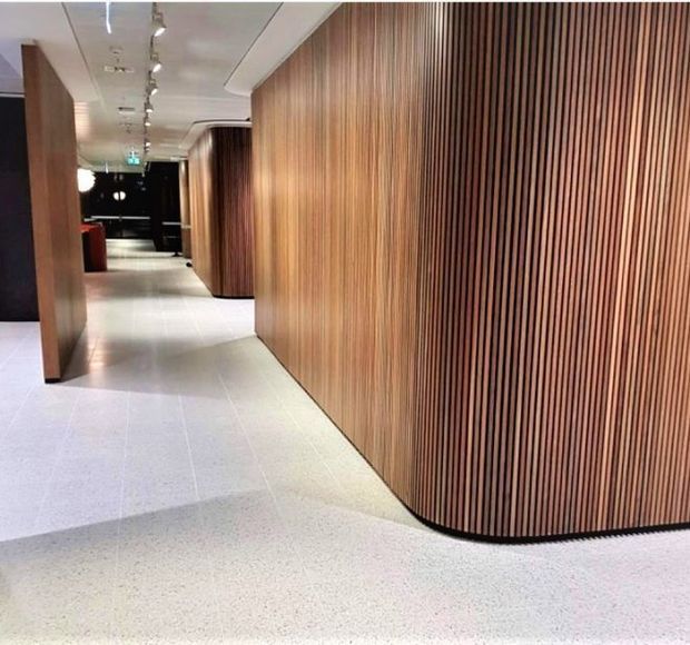 A contemporary commercial interior corridor featuring curved, floor-to-ceiling vertical timber slat wall panelling, pale terrazzo flooring, recessed ceiling details, and track lighting. The streamlined curved joinery creates a refined, modern passage leading toward a darker reception or lounge area.