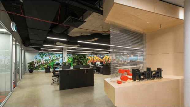 Contemporary open-plan commercial office reception and workspace. A pale timber reception desk with a prominent red sculptural logo sits in the foreground, while black workstations and office chairs occupy the open area behind it. Full-height glazing lines the left side, and a transparent textured partition separates the reception from the work area. The ceiling has exposed dark services, linear lighting and warm timber acoustic panels, with colourful botanical artwork and planting adding visual interest.