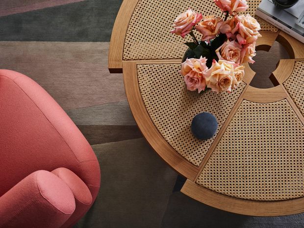 Overhead view of a richly styled seating vignette featuring a round timber coffee table with segmented woven cane insets, a coral-pink upholstered armchair, a muted geometric rug, a flower arrangement, and a dark decorative object. The composition has a warm, refined, mid-century-inspired character.