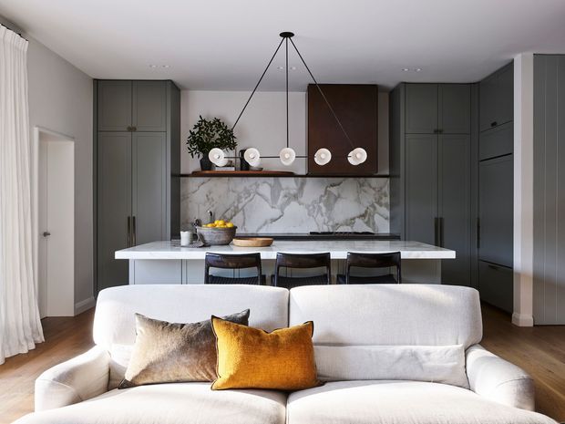 A refined open-plan living room with a contemporary kitchen behind a large white island. The room features dark grey full-height cabinetry, a dramatic white-and-grey veined marble backsplash, timber flooring, a sculptural linear pendant light, and a plush cream sofa with warm ochre and neutral cushions in the foreground.
