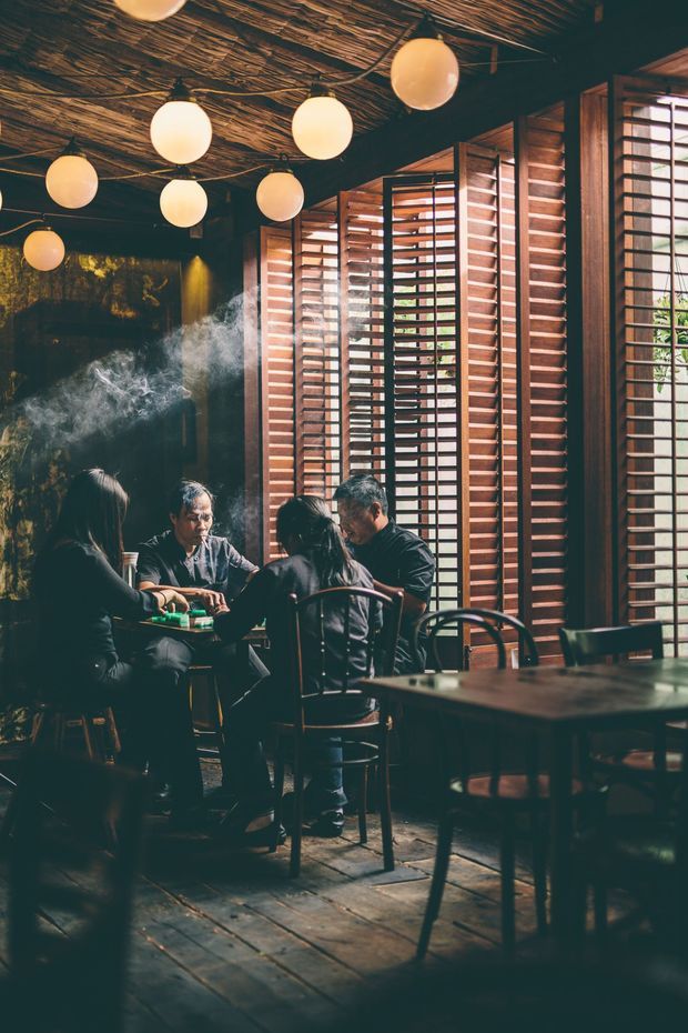 A dimly lit dining area in a restaurant or hospitality venue, with several diners seated at dark wooden tables. The space features warm globe pendant lights, a textured woven or branch-like ceiling, dark painted walls, timber plank flooring, and prominent timber horizontal louvres or shutters filtering daylight along the windows. The atmosphere is intimate, warm, and moody.