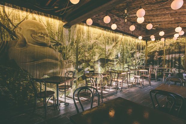 A dimly lit restaurant or café dining area with numerous small round tables and bentwood-style chairs. Large wall imagery and reflective glazing depict a lush forest scene, while a prominent Buddha-like figure appears at the left. The ceiling features exposed rustic timber branches or beams and a cluster of warm globe pendant lights, creating an atmospheric, immersive interior.