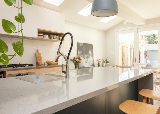 Bright contemporary kitchen viewed from the island, featuring a large pale stone island benchtop with integrated sink and curved black tap, dark lower cabinetry, light timber cabinetry, open shelving, gas cooktop, skylights, and glass doors opening to an outdoor area. A large pendant light and indoor plants add visual interest.