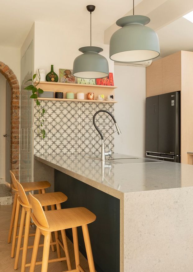 A warm, contemporary kitchen centred on a large pale stone island with a dark base, integrated sink and curved gooseneck tap. Three timber bar stools line the island. The kitchen features a patterned grey and white tiled splashback, open display shelving, light timber cabinetry, a dark double-door refrigerator, oversized muted green pendant lights, and an exposed brick arched opening at the left.