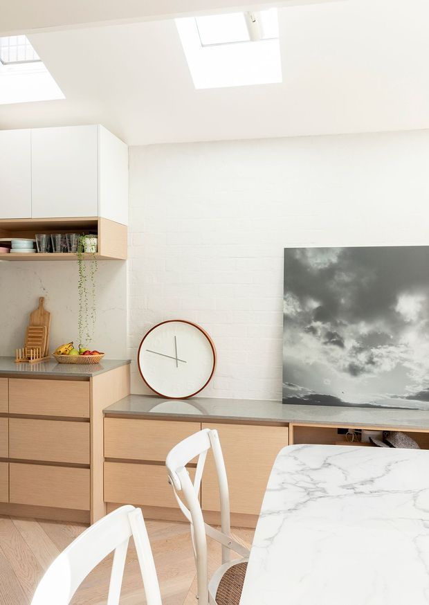 Bright, compact open-plan kitchen and dining area with white cabinetry, light timber drawer fronts, a grey benchtop, white-painted brick walls and skylights. A marble-look dining table and white chairs occupy the foreground, while a large monochrome artwork and round wall clock add visual interest.