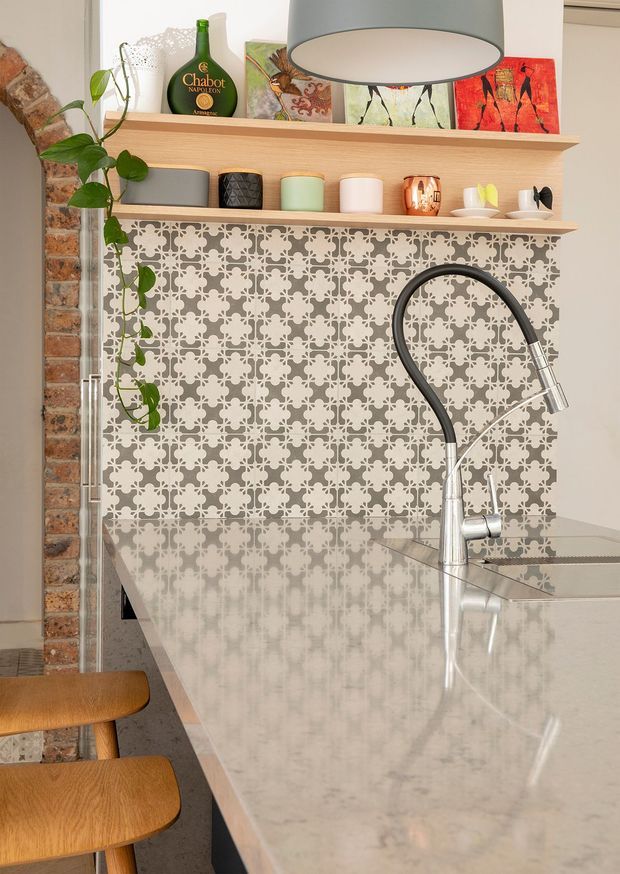Close-up view of a characterful kitchen featuring a large polished light-grey stone island with an integrated sink and tall black-and-chrome gooseneck tap. The rear wall has a decorative repeating grey-and-white patterned tile splashback, open timber shelves displaying cookware and decorative objects, exposed brickwork to the side, and a large grey pendant light overhead. Timber bar stools and trailing greenery add warmth and contrast.