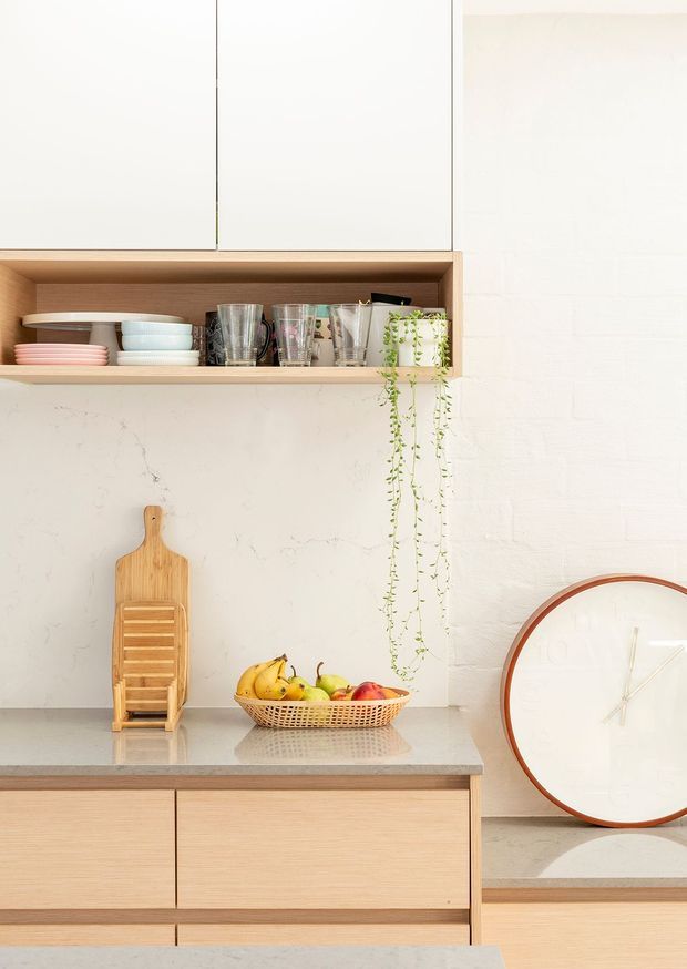 Bright, softly styled kitchen vignette with pale timber cabinetry, a light stone-look benchtop and backsplash, open timber shelving displaying crockery and glassware, a trailing plant, fruit bowl and oversized round clock. The composition is warm, minimal and natural, with clean lines and restrained decor.