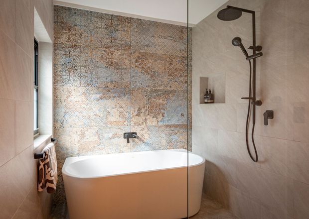 A refined contemporary bathroom featuring a freestanding white bathtub beside a frameless glass shower enclosure. The shower walls are clad in large pale stone-look tiles, while the wall behind the bath has a prominent blue-grey and warm beige patterned feature tile. Dark bronze or black shower fittings, a recessed shelf, and a small window complete the warm, textural scheme.
